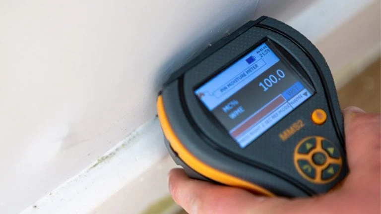 A moisture meter being held against a wall with signs of damp. The reading shows 100% saturation.