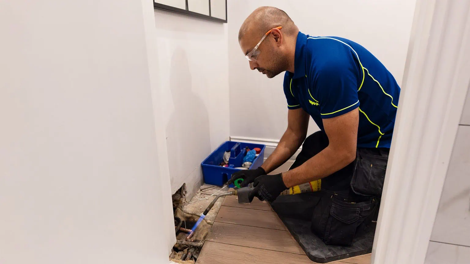 An Aspect plumber uses a gas torch to repair a leak in a copper pipe exposed beneath a bathroom floor.